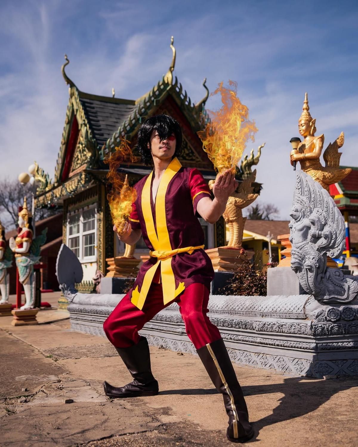Outdoor customer cosplay shot by @jcw.cosplay at buddhist temple, Zuko in firebending combat stance with flame effects on both hands, maroon top with gold trim, red pants, black curled boots, scar makeup over left eye
