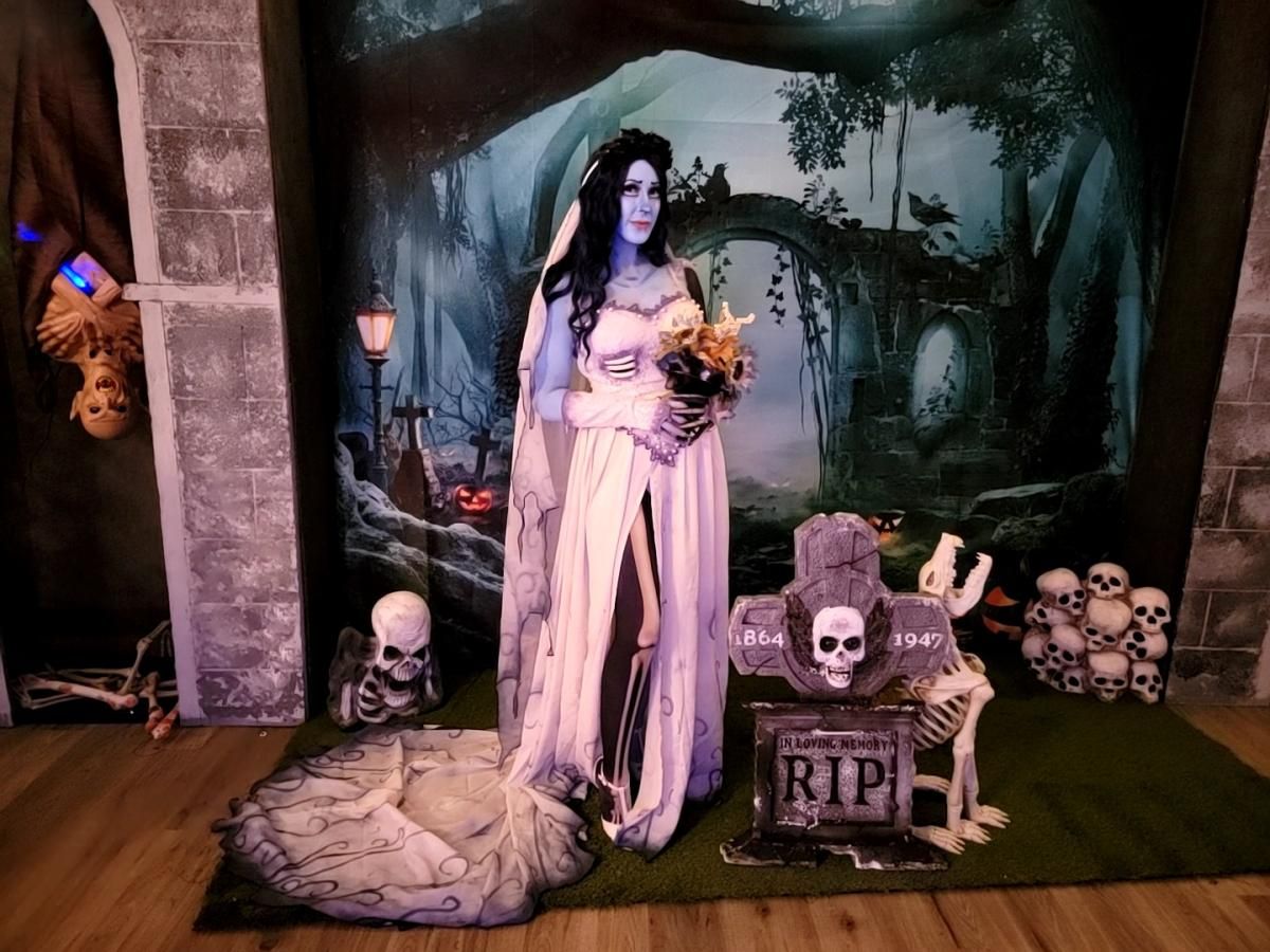 Indoor themed customer shot by @Alice in front of tombstone & skeleton Halloween backdrop, full sheer ghost wedding gown with trailing train, lace long gloves and veil, holding dried flower bouquet with spooky graveyard props surrounding the scene