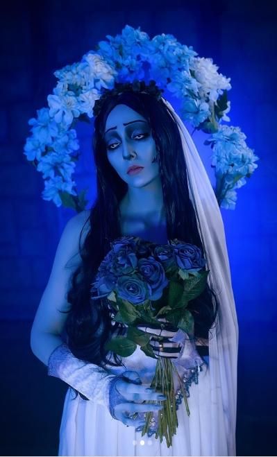 Close half-body studio portrait under soft blue-purple lighting from @bellatrixaiden, oversized blue-white floral halo headpiece on top of long gradient wig, lace skeleton gloves gripping matching blue rose dead flower bouquet