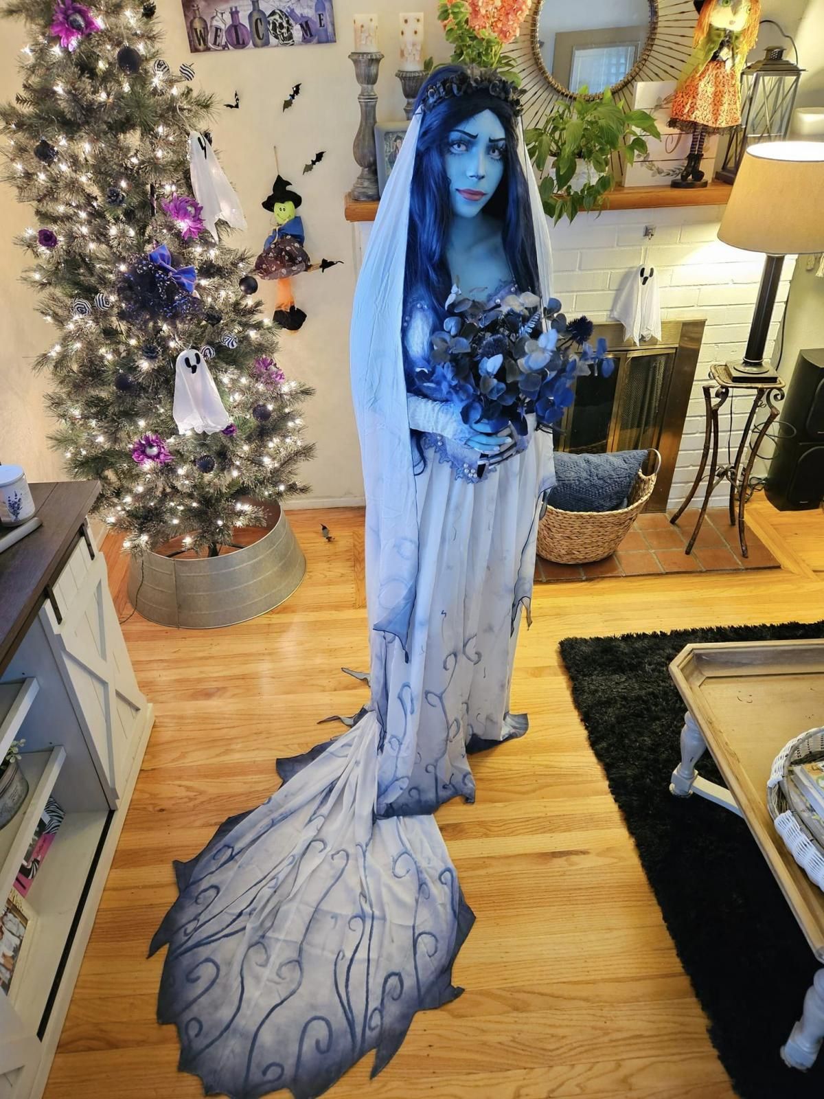 Front full-body indoor shot of @Cassi in complete Emily cos outfit, holding matching dark blue dried flower bouquet, dark floral wreath headpiece, full gradient veil, layered sheer wedding gown with long jagged train, holiday decorated fireplace and tree backdrop
