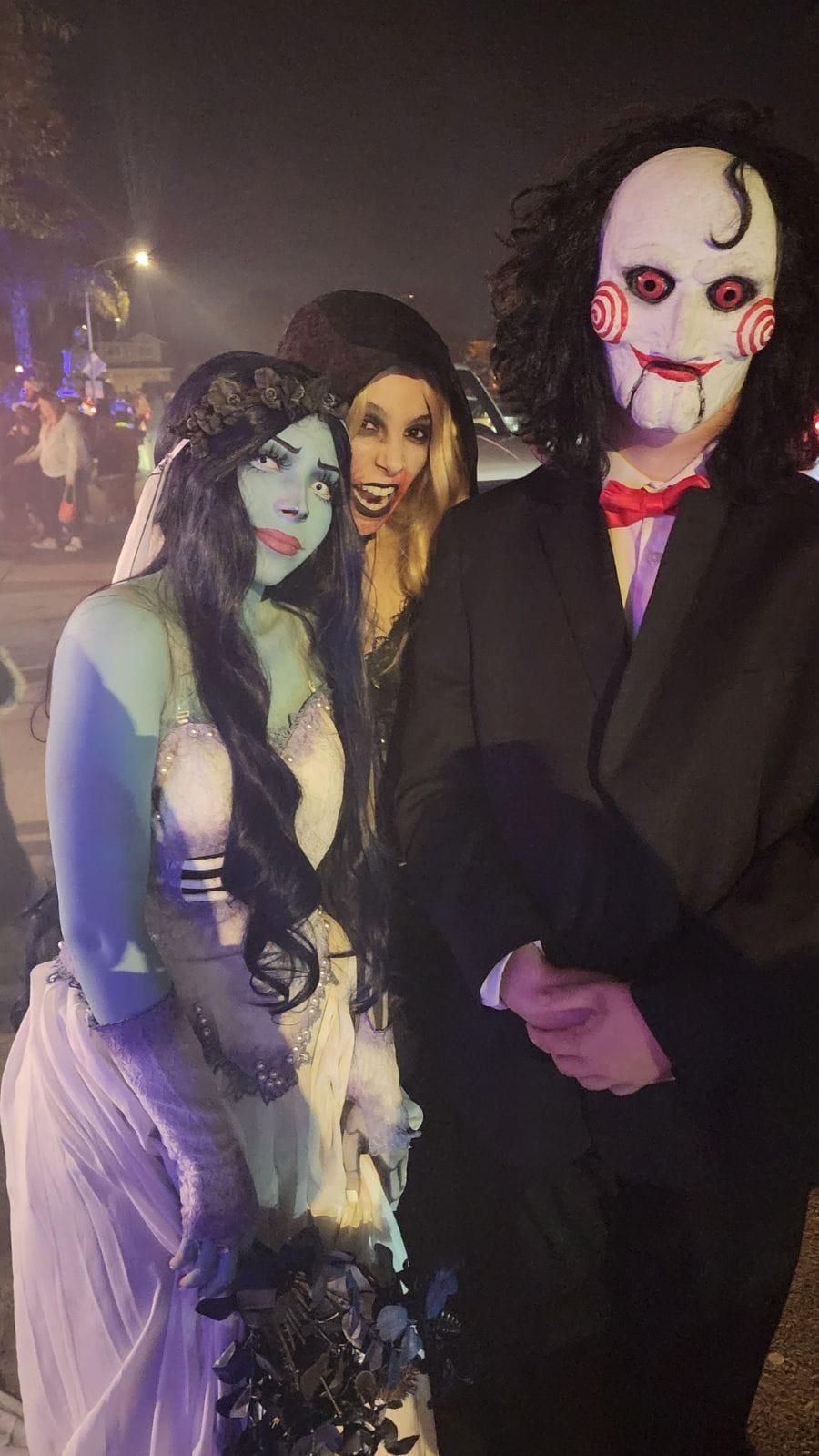Night outdoor Halloween street group portrait shot by buyer @Cassi, full Emily ghost bridal cos set including gradient sheer wedding gown, dark floral crown, long lace skeleton gloves, long dark blue gradient wig, holding dark blue dead flower bouquet, posing alongside other horror themed cosplayers under street lamp ambient lighting