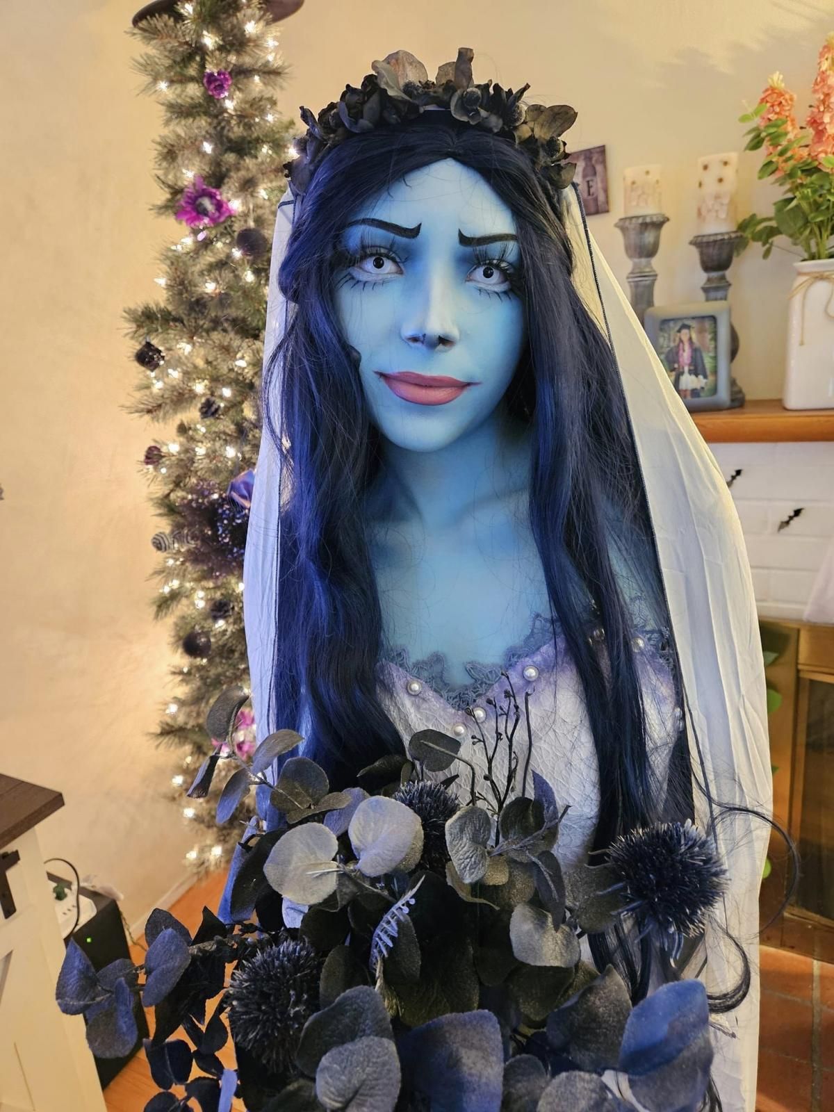 Close-up indoor portrait of @Cassi focusing on dark dried flower halo headpiece and matching dark blue eucalyptus bouquet, long wavy dark gradient wig framing blue character makeup, pearl lace trim on corset neckline clearly visible