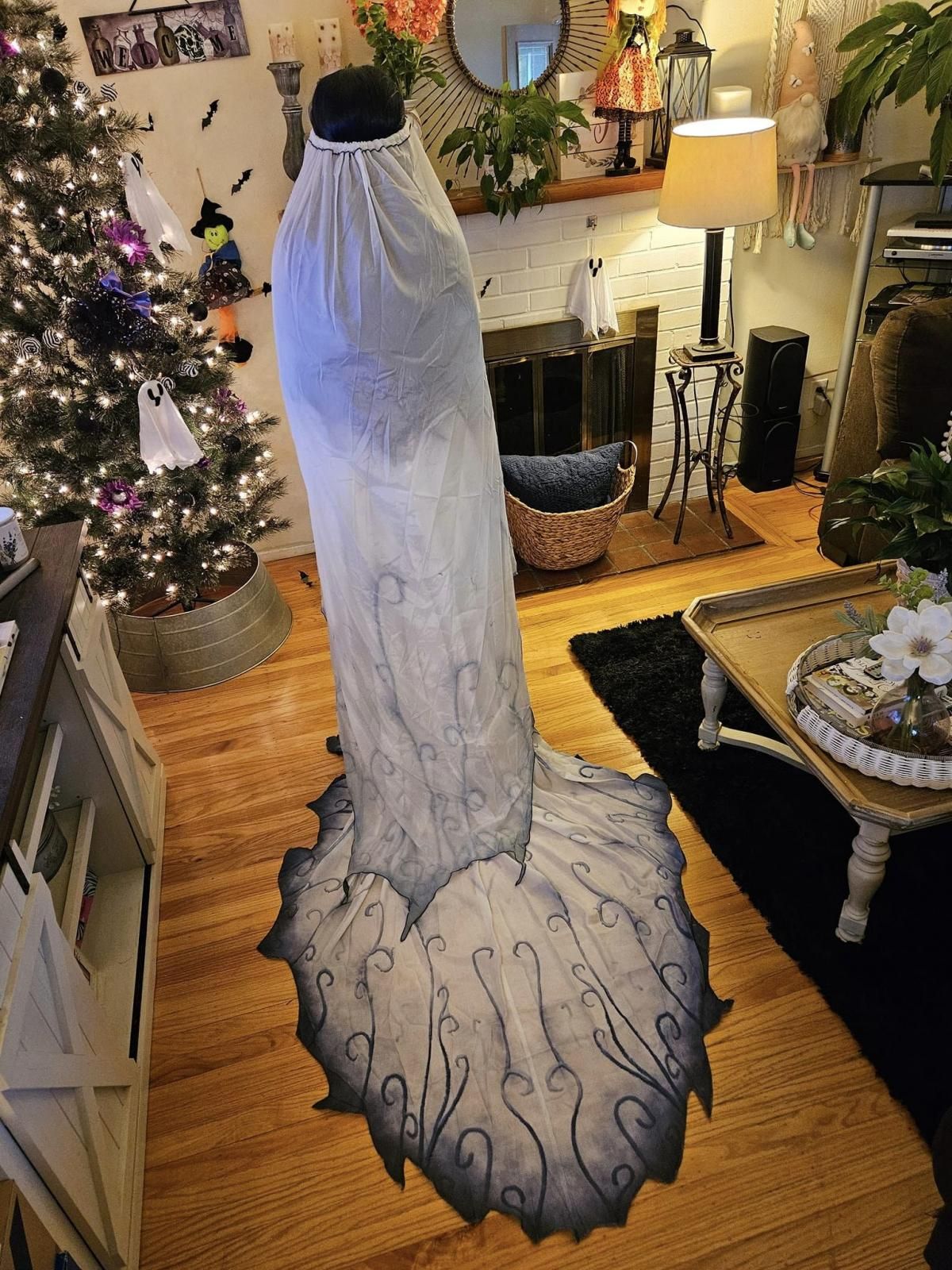 Full back trail indoor shot of @Cassi standing in Halloween decorated living room, complete view of irregular jagged chiffon train covered in continuous dark vine swirl print, long gradient veil draping all the way down the spine, Christmas tree with ghost ornaments as background
