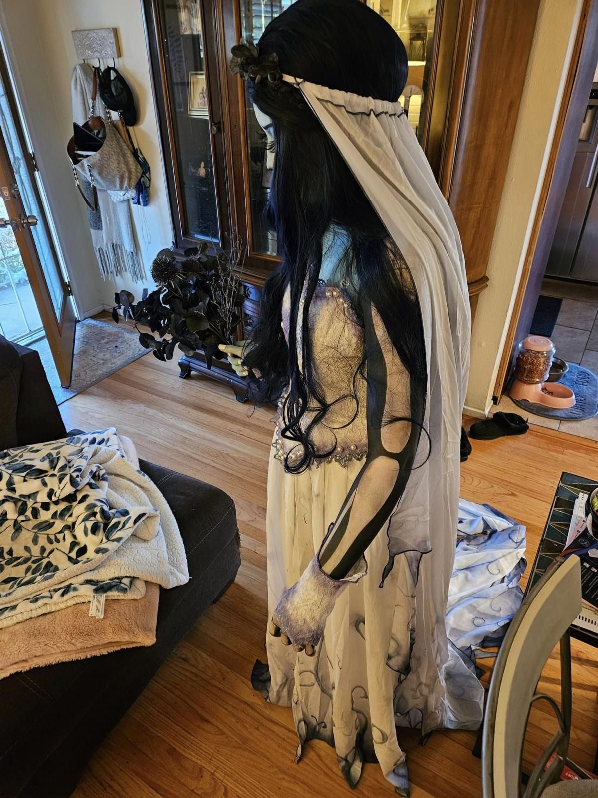 Indoor home side profile shot of @Cassi, focus on printed hollow skeleton arm detail through thin sheer gown fabric, full layered long veil, dark floral hair wreath and pearl trimmed corset bodice, warm wood floor indoor background