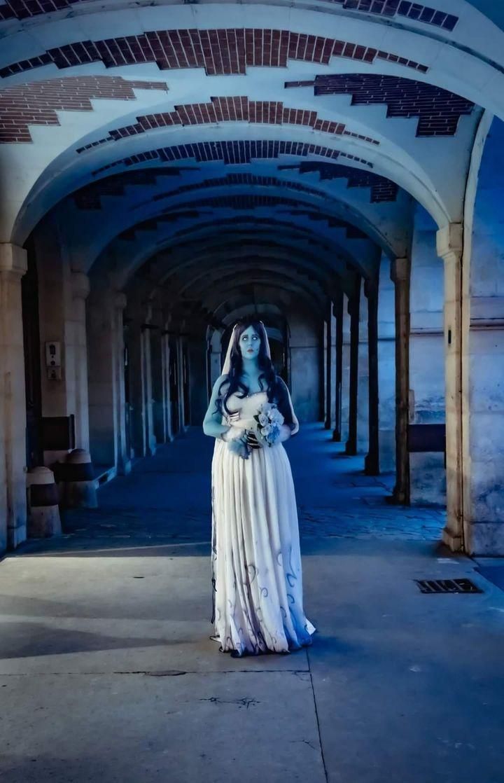 Wide full-body distant outdoor shot of @Heiko no Sumi standing in misty cool blue toned brick arcade, full floor-length gradient ghost wedding gown with natural train spread across stone pavement, holding light blue rose bouquet centered in frame