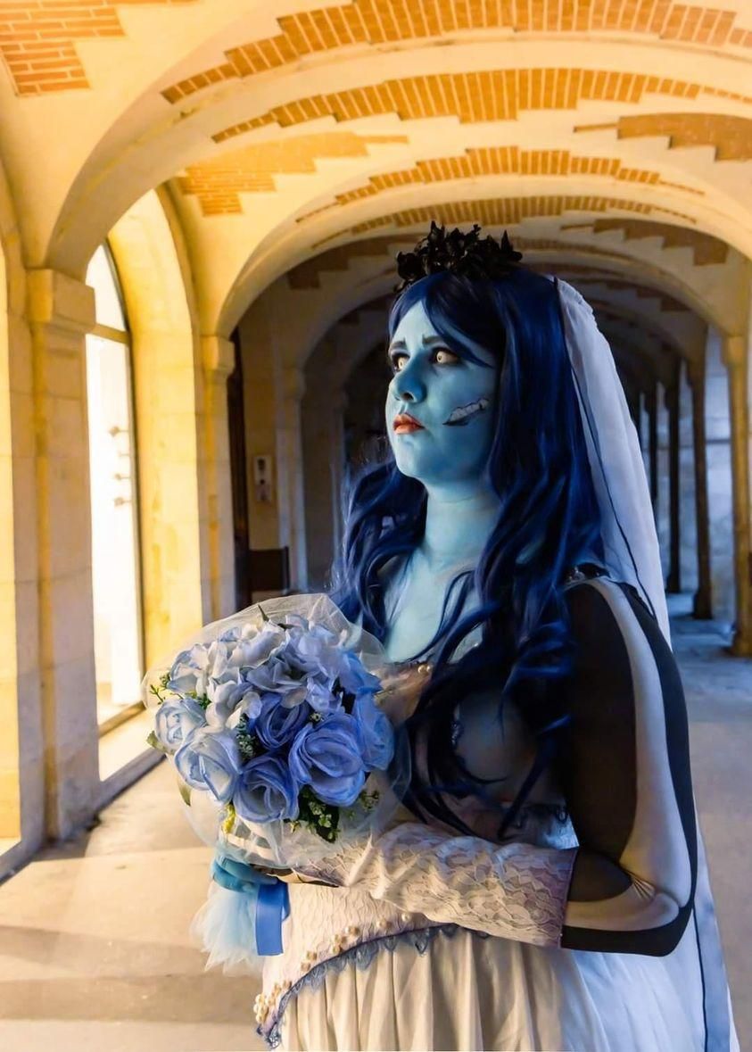 Warm stone arcade corridor outdoor half-body shot of @Heiko no Sumi, holding soft pale blue rose bouquet, dark floral hair crown and long sheer veil, hollow printed skeleton sleeve detail visible on upper arm, arched brick building architectural backdrop