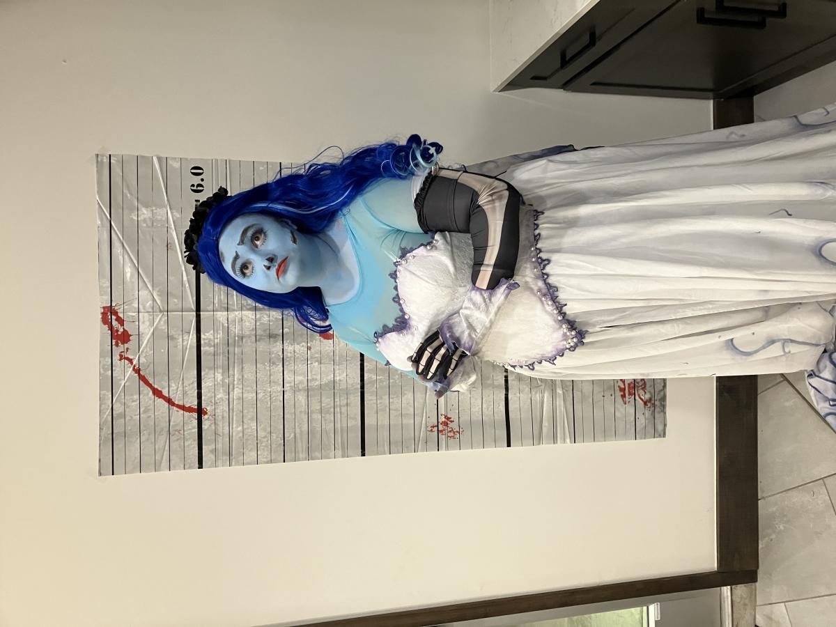 Indoor full-body shot of buyer @Jillian in complete Emily ghost bridal outfit, blue gradient long wig, dark floral head wreath, pearl lace corset bodice, sheer mesh inner top, printed skeleton long gloves and full swirling vine print chiffon skirt, blood splatter mugshot backdrop as decoration
