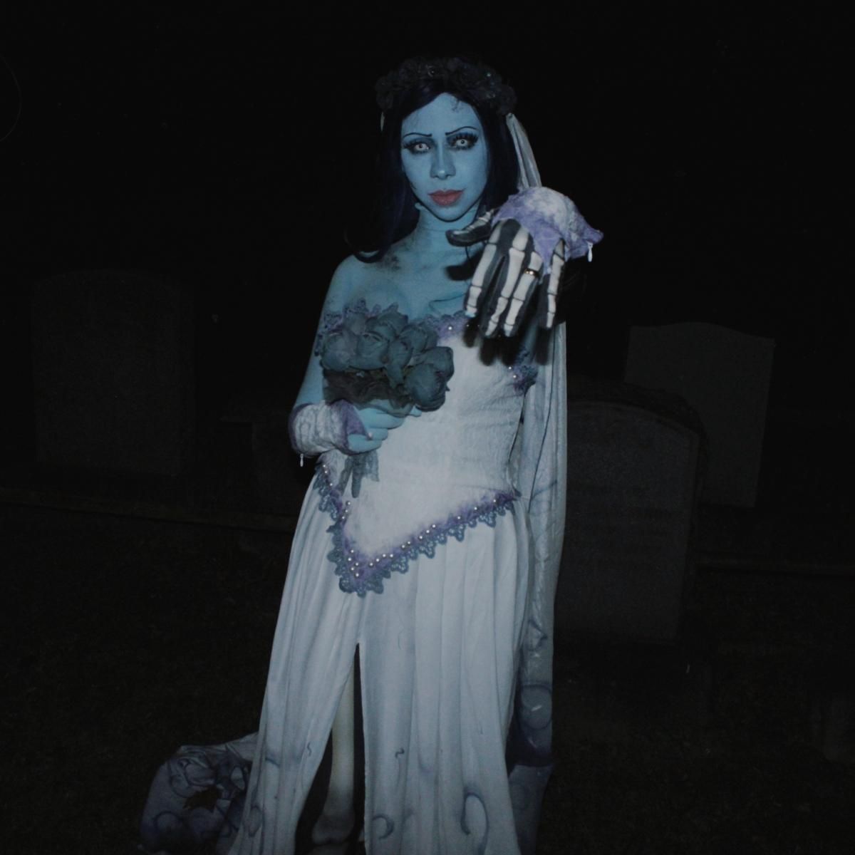 Night cemetery front close half-body shot of @Kaysie, focus on skeleton segmented print long gloves and matching dark blue rose bouquet, pearl lace trim on corset hem, dark floral crown above forehead, tombstones dim background