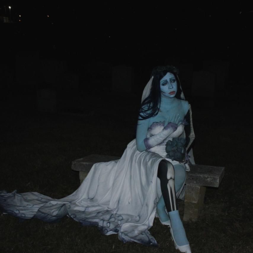 Dark night cemetery outdoor shot of @Kaysie sitting on stone bench, long flowing gradient chiffon train spread across grass ground, printed skeleton leg detail visible on thigh, holding dark blue dried flower bouquet, tombstones dim night scenery background