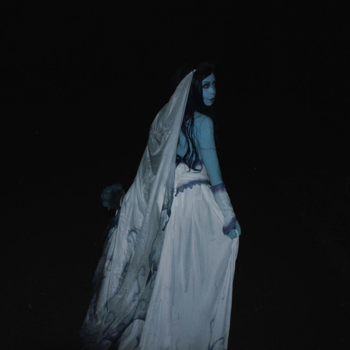 Back view full silhouette night shot of @Kaysie against total black dark background, full length gradient swirling print chiffon train, wide layered sheer veil trailing down spine, hollow printed skeleton arm detail visible on outer sleeve