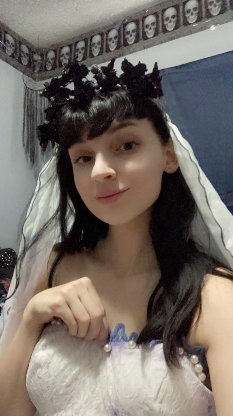 Indoor home close-up selfie of @Raven focusing on matching dark dried flower wreath headpiece and long sheer veil, pearl lace corset neckline detail clearly visible, skull patterned wall border interior background