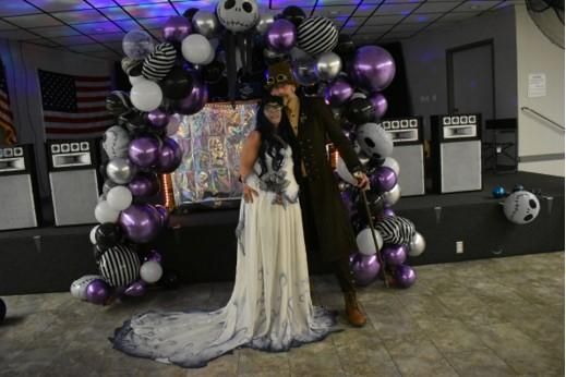 Indoor event stage full-body duo shot of @Speedy wearing full Emily ghost bridal gown with long gradient train, paired with dark vintage long coat cosplayer, purple silver skull themed balloon arch stage backdrop, hall activity venue scene