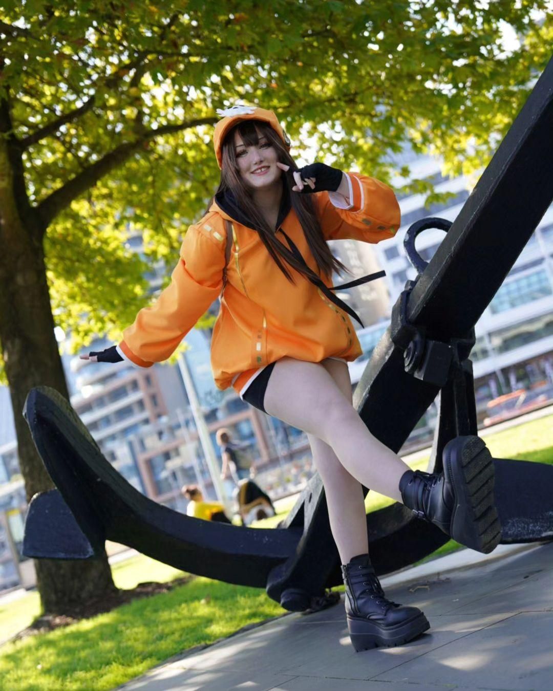 Full outdoor customer cos photo of Guilty Gear Strive May by @Kiri, posing beside huge ship anchor prop, complete orange hoodie, shorts, fingerless gloves and platform boots