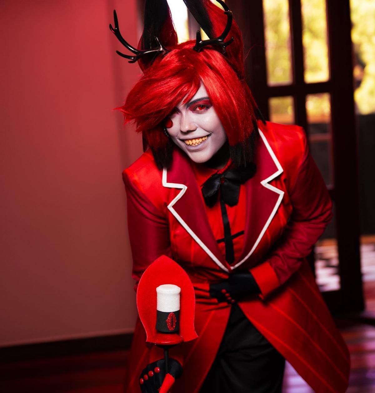 Indoor warm red tone half-body shot of buyer @arcee.cos leaning forward, full Alastor cos set including red pinstripe long coat with white lapel piping, black bow tie, cross printed inner shirt, claw gloves, red gradient wig, deer horn head accessories, holding signature radio staff prop, classic sharp-tooth character makeup
