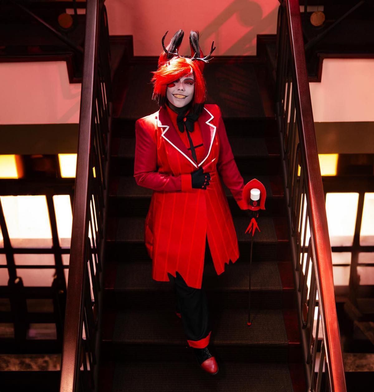 Full-body shot of @arcee.cos walking down dark vintage staircase, complete red pinstripe long coat, black cuffed trousers, red leather shoes, deer horn ear accessories, claw gloves gripping radio staff, red ambient lighting strengthens retro radio demon atmosphere