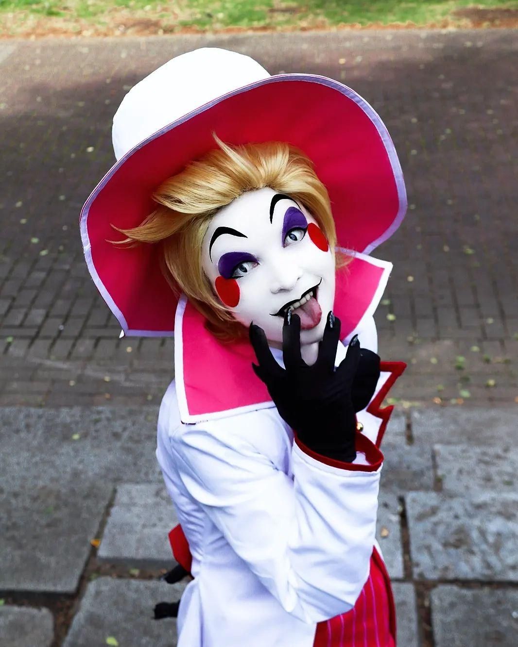 Outdoor pavement half-body shot of buyer @cantarella_creative, full Lucifer cos outfit including white red-trim tailcoat, red striped waistcoat, black bow tie, classic white tall top hat with red brim, black claw gloves, blonde styled wig, full character clown stage makeup, playful tongue-out posing