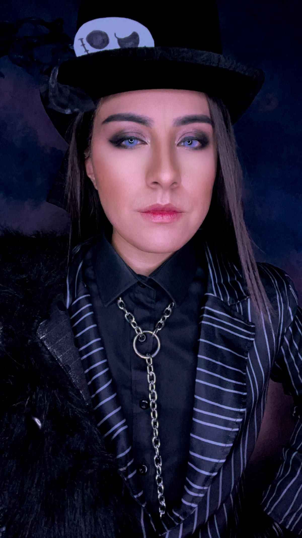 Indoor close half-body portrait from @Angel, styled with Jack pinstripe blazer, skull print top hat and punk metal chain accessory, soft smoky eye makeup to match spooky goth aesthetic