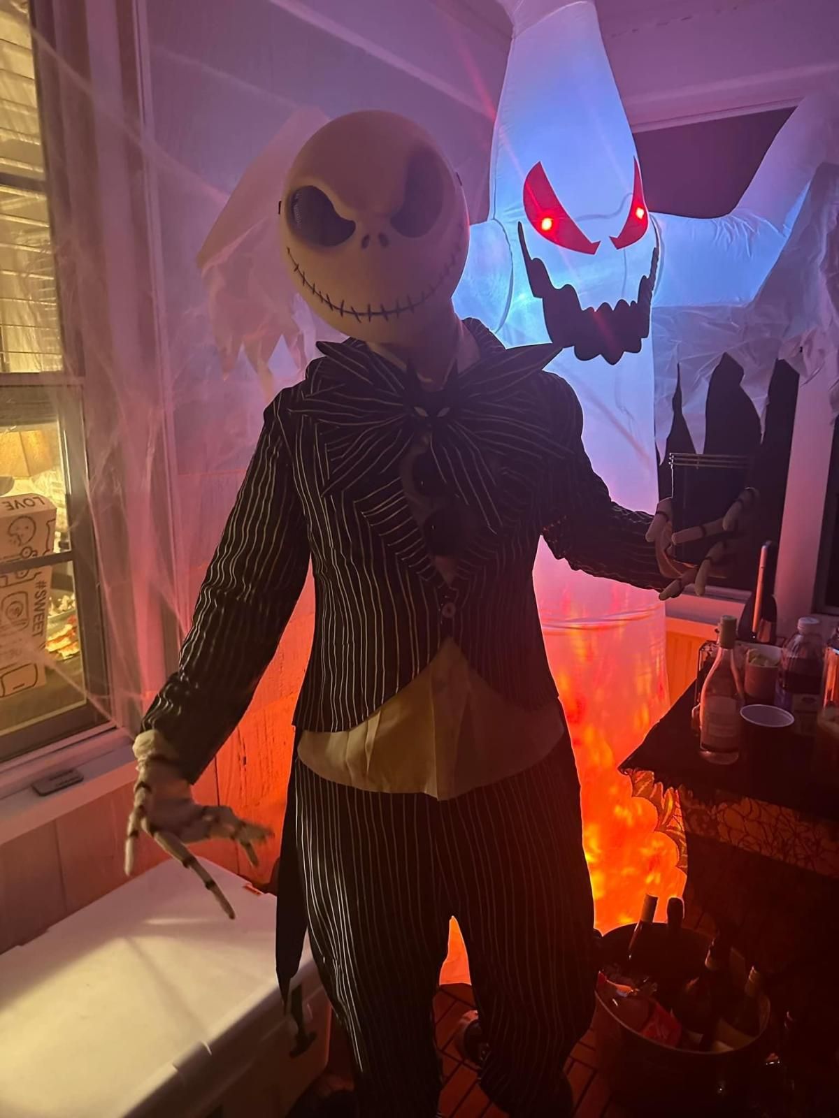Halloween indoor party customer shot by @Cpitt, wearing full Jack suit, latex skull mask and bony finger gloves, giant inflatable ghost decoration as backdrop with festive drink table scene