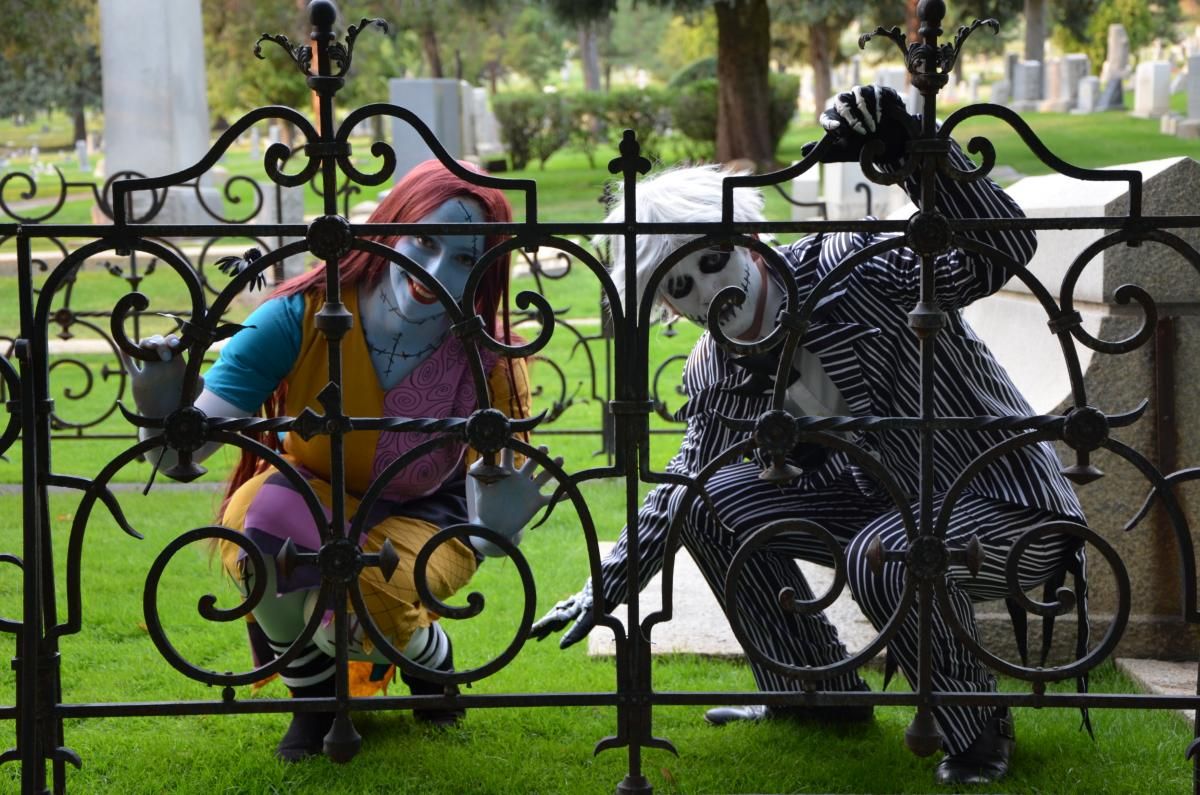 Outdoor cemetery full-body customer shot by @ammotc, Jack Skellington in classic black-white pinstripe suit paired with Sally patchwork costume, posing behind wrought iron fence for spooky Halloween themed photos