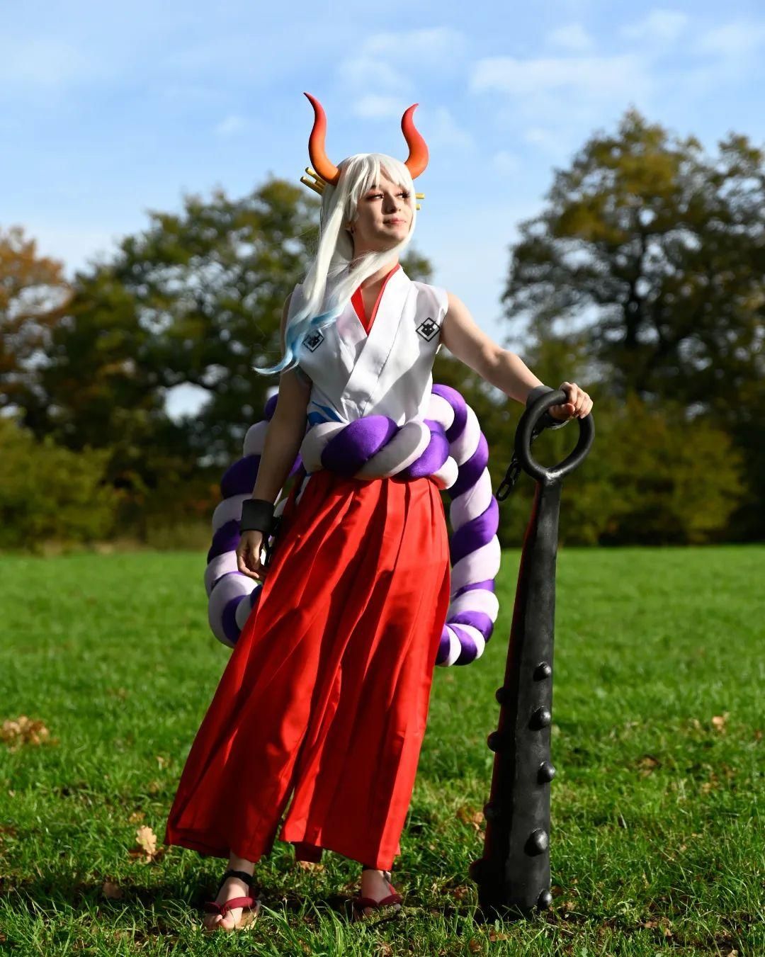 Full‑body outdoor cosplay photo by @corvus.cosplay for One Piece Yamato on green meadow, leaning on large kanabo club prop, horns and waist rope decoration.