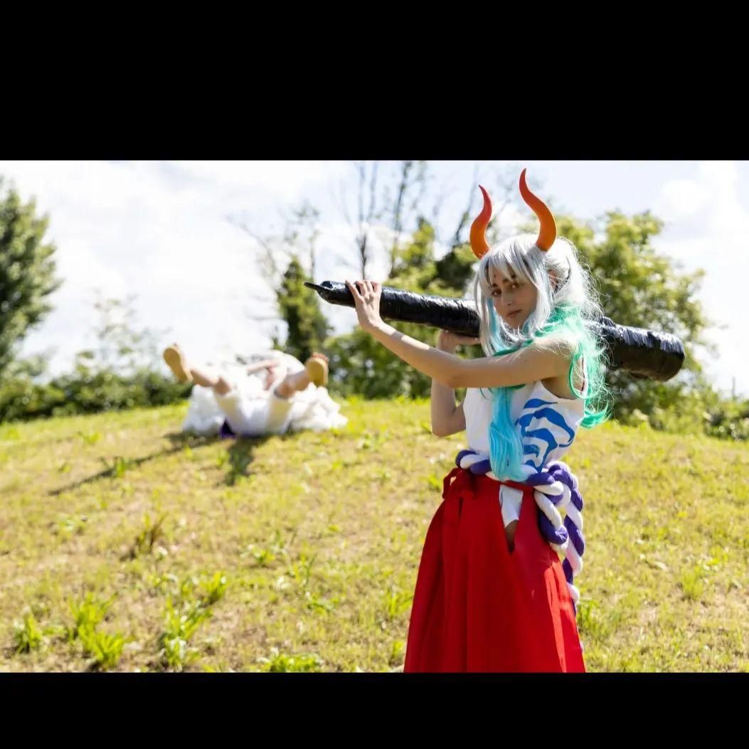 Outdoor grassland action cosplay shot by @Aury as One Piece Yamato, holding large club prop across shoulder, red hakama and gradient styled wig.