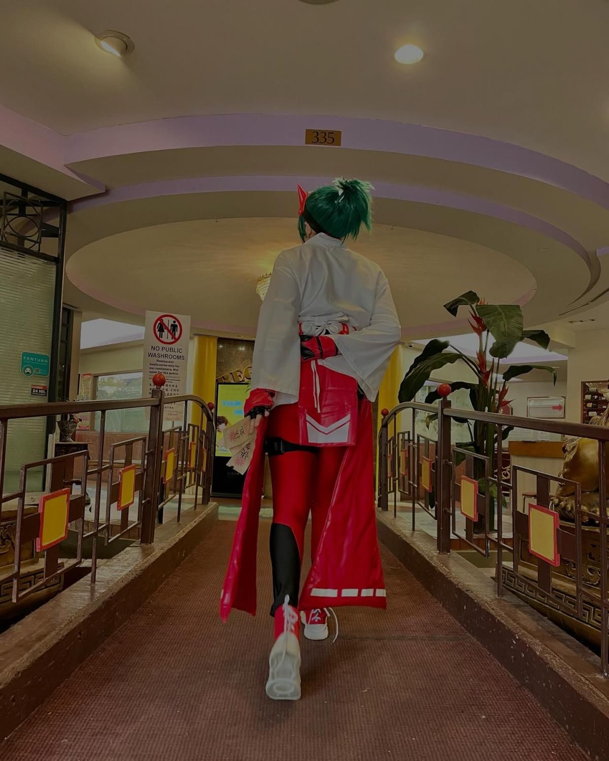 Back view full‑body customer shot by @stephanieschaerer inside building corridor, kiriko cosplay costume showing obi, long red tail pieces, teal ponytail wig and cos boots