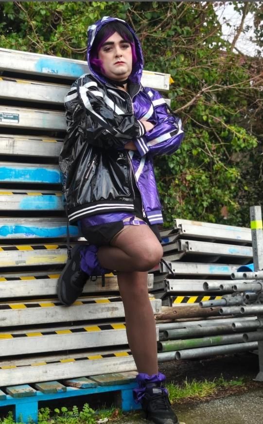 Full body outdoor customer photo by @aquameloncosplay, complete Reina cos set including shiny jacket, black shorts, thigh harness stockings, purple ankle shoes, posing on stacked metal pallets