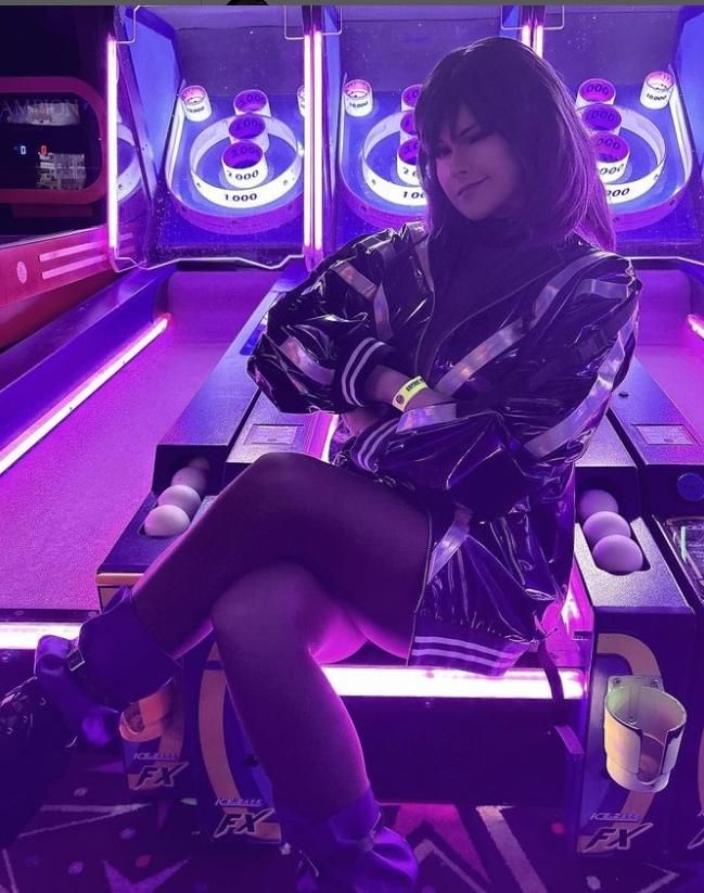 Atmospheric seated customer shot by @v0dch4n inside neon arcade game room, full Reina cos glossy jacket, sheer stockings, purple ankle boots, sitting on skeeball game machine with purple neon lighting