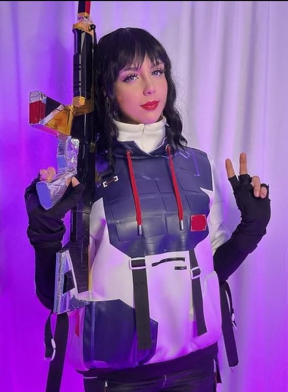 Studio shot under purple ambient lighting of customer @susbaddie in full ISO top, gripping large game rifle prop against shoulder, grey-accented fingerless gloves and layered hoodie vest structure fully visible